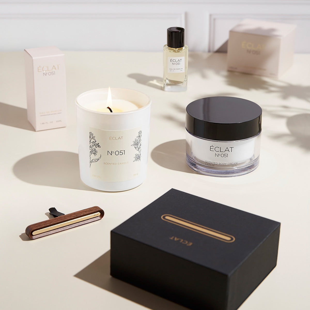 ÉCLAT No. 051 women's fragrance set displayed on a light surface, featuring a lit scented candle, a jar of scented body cream, a bottle of Eau de Parfum, a black and gold car scent clip, and matching product packaging. The arrangement exudes elegance and femininity.