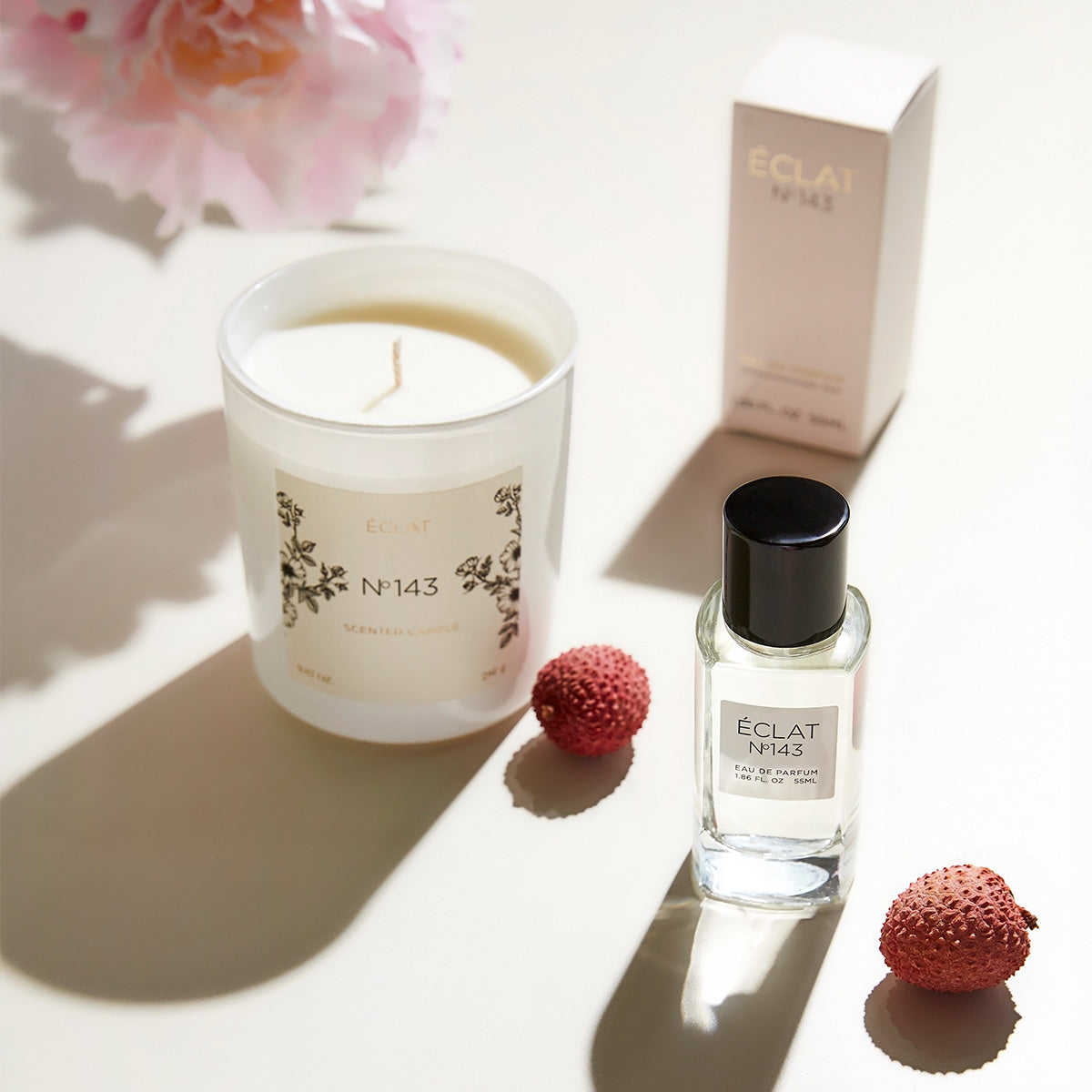 ÉCLAT Clean Rose perfume and ÉCLAT No. 143 scented candle set, displayed with lychee fruits on a light surface. The perfume bottle features a black cap and white label, while the matching candle is housed in frosted glass with floral design elements. A matching perfume box is in the background.