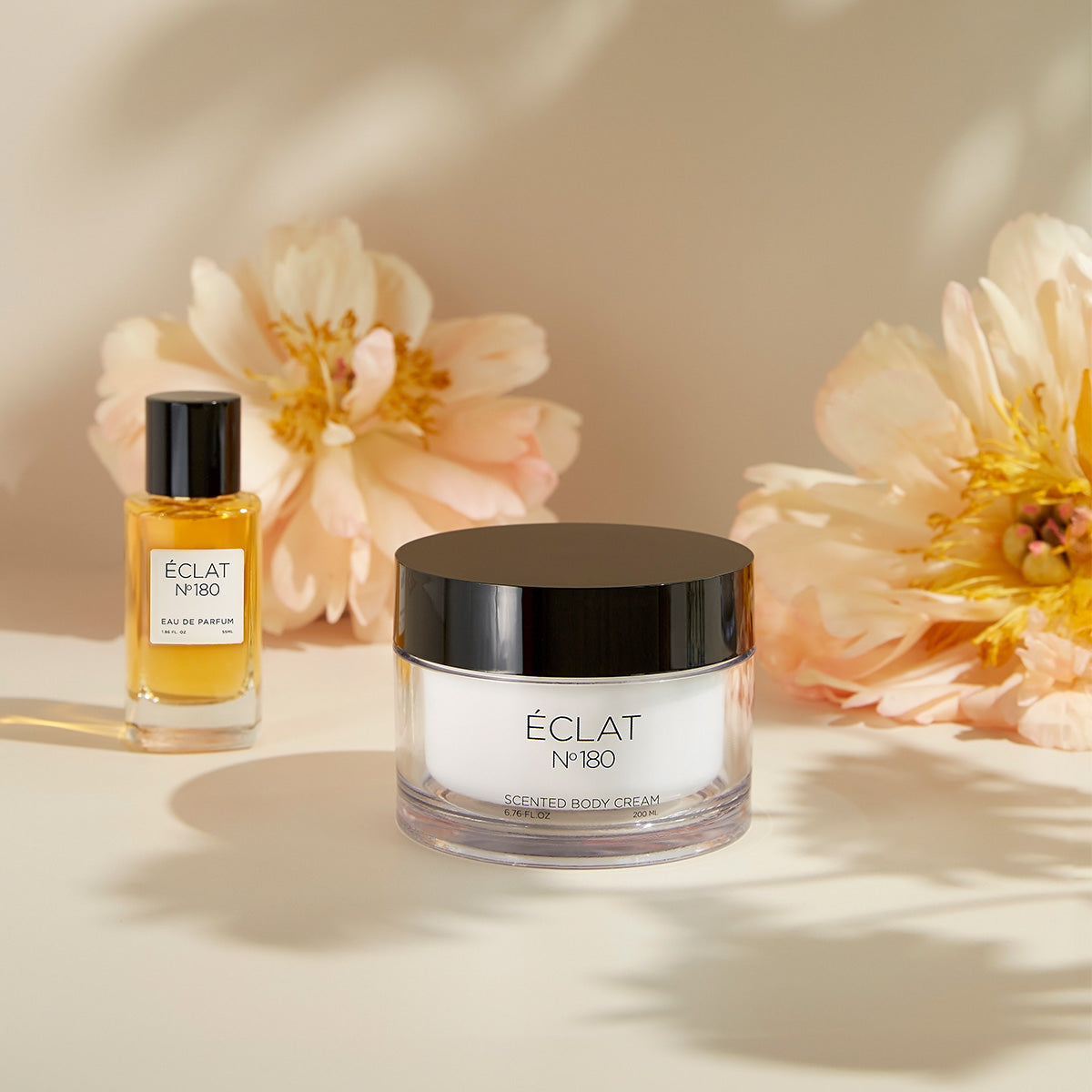 ÉCLAT No. 180 women's perfume and scented body cream displayed among soft, blooming peonies. The warm-toned perfume bottle and the elegant cream jar with a black lid are bathed in gentle natural light, highlighting the feminine and floral character of the fragrance duo.