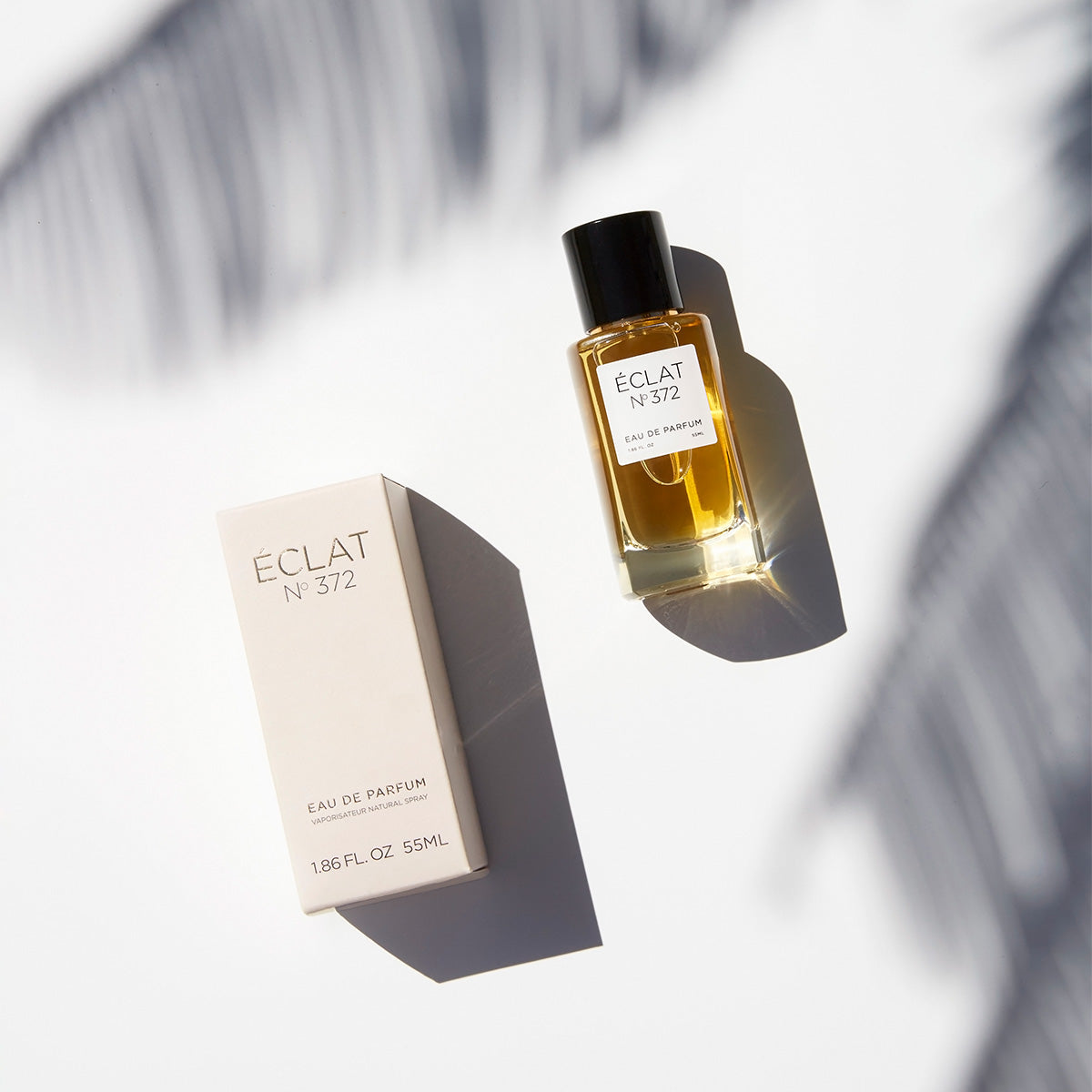 ÉCLAT No. 372 women's perfume captured in bright, natural sunlight. The amber-colored fragrance is housed in a sleek glass bottle with a black cap, lying beside its matching light beige packaging. Palm leaf shadows play across the white surface, evoking a summery, elegant vibe.