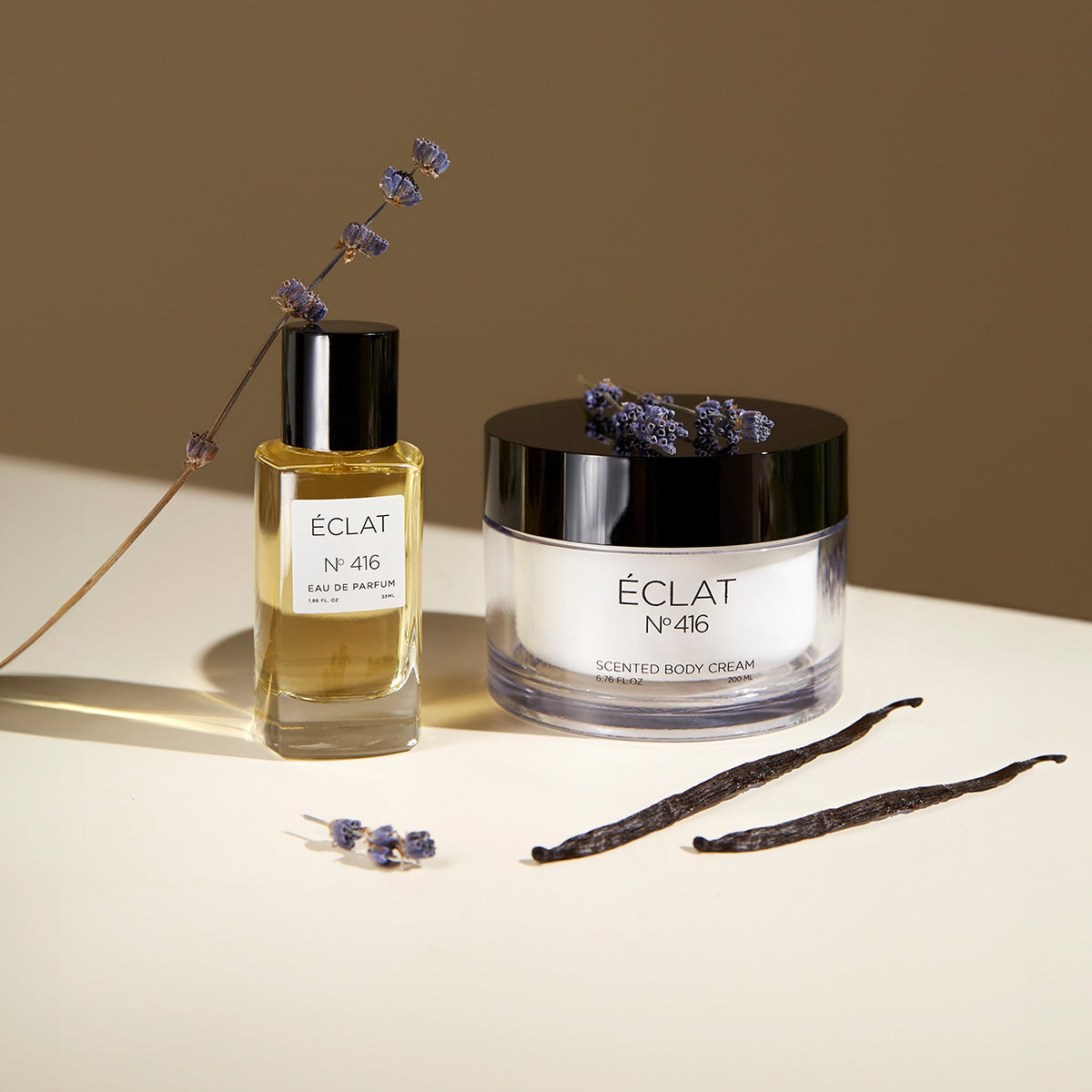 ÉCLAT No. 416 women's perfume and scented body cream set, elegantly arranged on a light surface. The golden-hued perfume is in a clear glass flacon, while the luxurious body cream comes in a sleek jar with a black lid. Lavender sprigs and vanilla pods are artfully placed, reflecting the fragrance’s floral and warm notes.