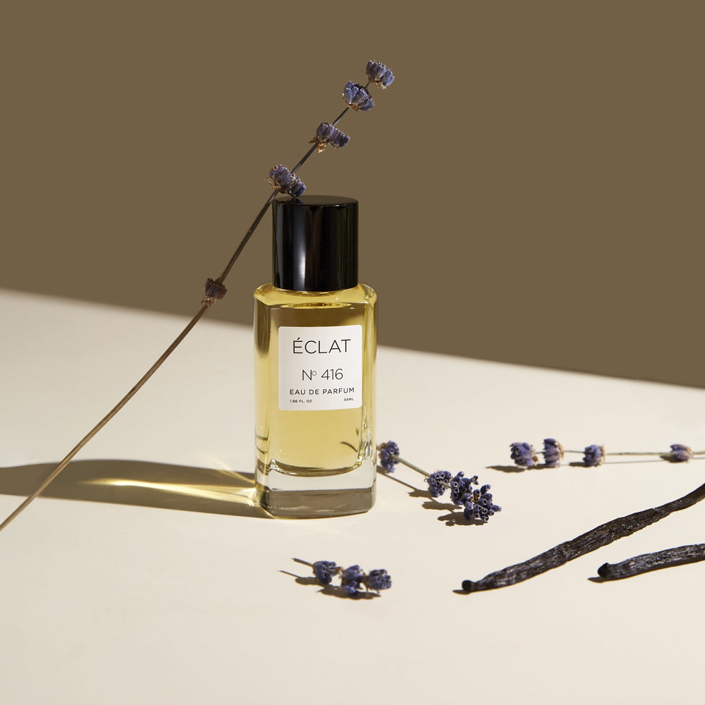 ÉCLAT No. 416 women's perfume bottle displayed on a light surface, surrounded by dried lavender sprigs and vanilla beans. The golden liquid in the clear glass flacon reflects the soft lighting, creating a warm and elegant composition.