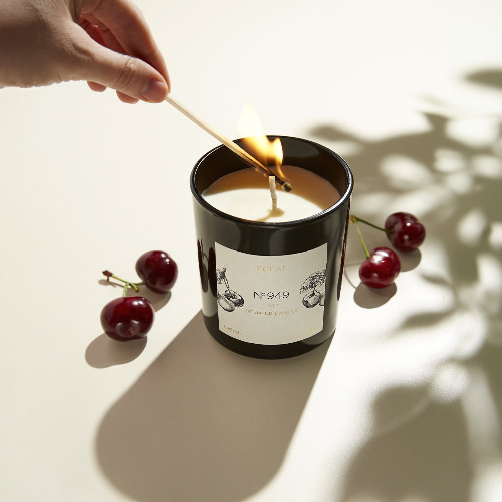 A black ÉCLAT scented candle labeled No. 949 is lit with a match, surrounded by fresh cherries on a light surface.