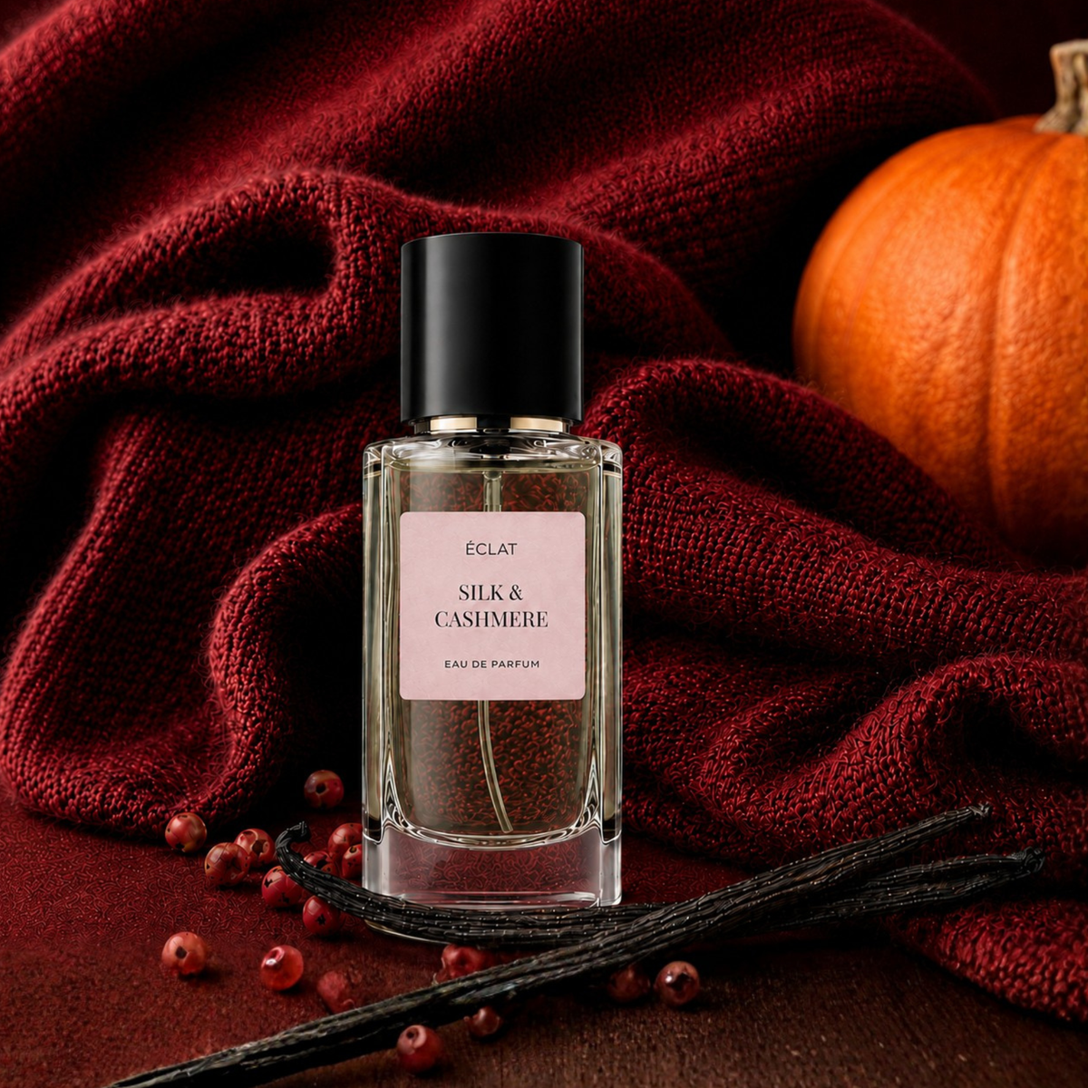 Un flacon en verre facetté de ÉCLAT Silk & Cashmere avec un bouchon noir, posé sur un fond de tissu rouge profond, entouré de gousses de vanille, de baies et d'une citrouille.