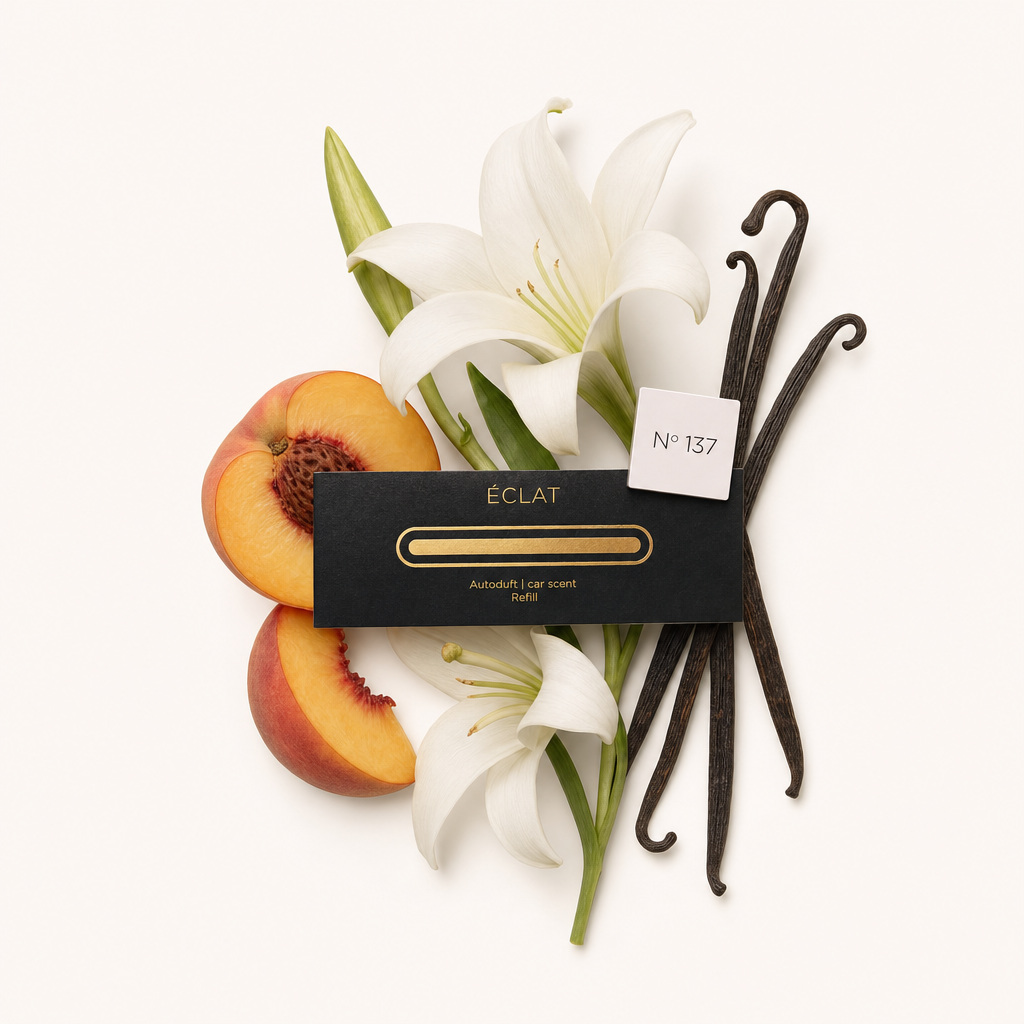 An arrangement of vanilla pods, white lilies, and peach halves surrounds a black ÉCLAT car fragrance cap labeled No° 137.