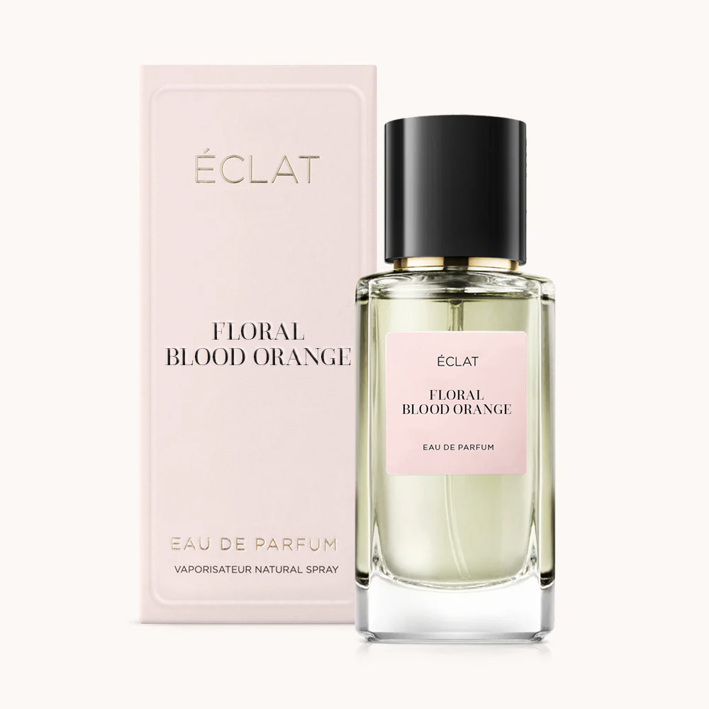 Un frasco de vidrio facetado de ÉCLAT Floral Blood Orange con tapa negra al lado de una caja rosa pálido.