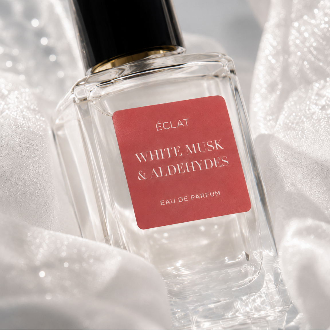 Un flacon en verre facetté de ÉCLAT White Musk & Aldehydes avec une étiquette rouge et un bouchon noir, entouré de tissu blanc délicat.