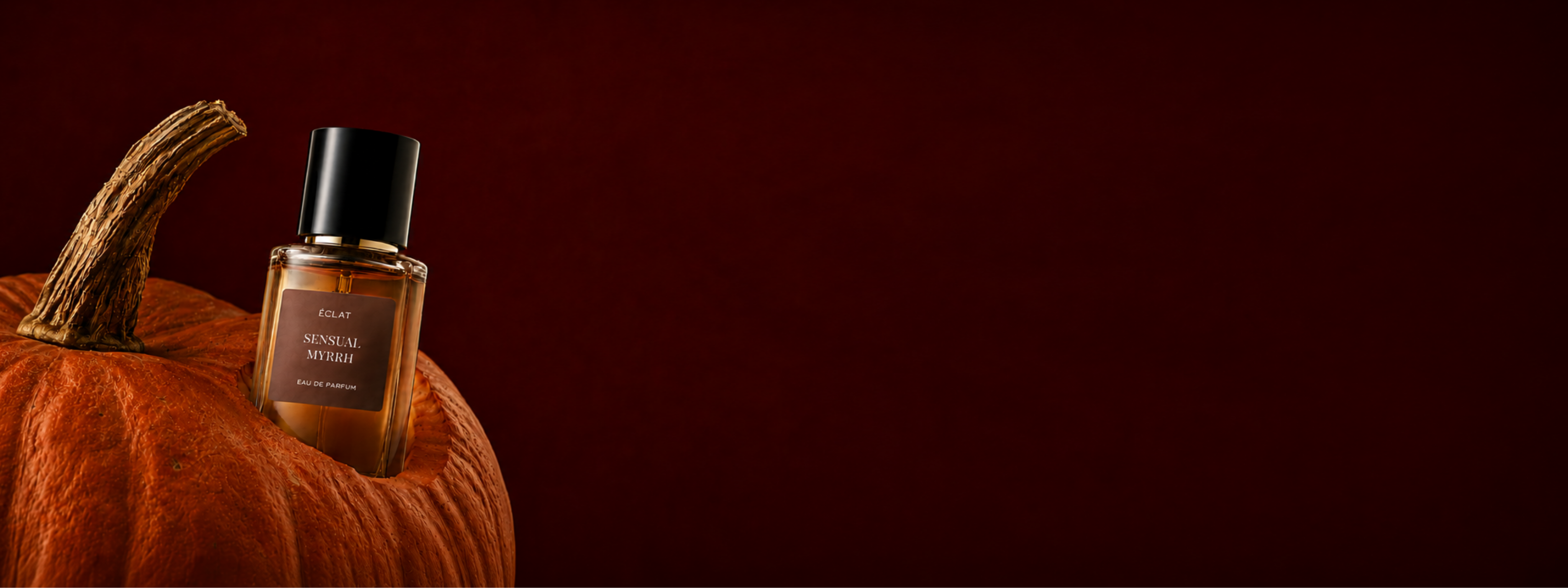 A faceted glass bottle of ÉCLAT Sensual Myrrh perfume is elegantly placed on a pumpkin against a rich, dark red background.