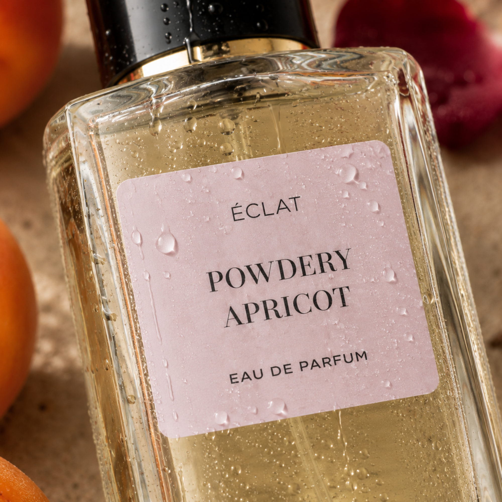 Flacon en verre facetté d'ÉCLAT Powdery Apricot avec étiquette rose pâle et bouchon noir, entouré d'abricot et de pétales rouges.