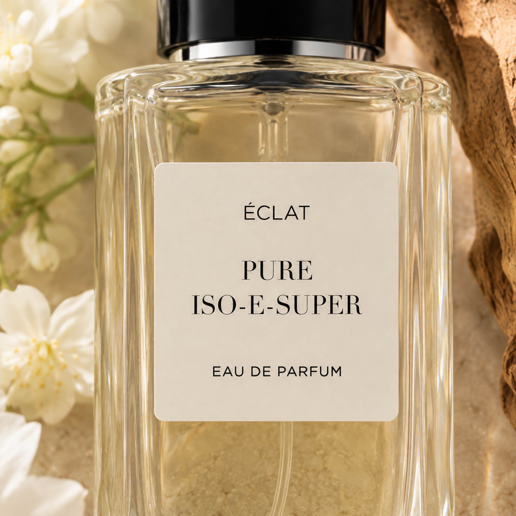Ένα φιάλη από γυαλί με έδρες του ÉCLAT Pure Iso-E-Super με μαύρο καπάκι, περιτριγυρισμένο από λευκά άνθη και υφή ξύλου.