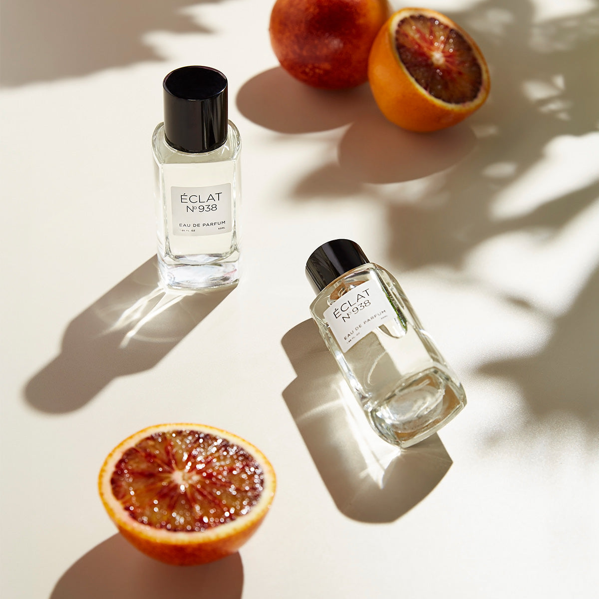 Two faceted glass bottles of ÉCLAT Ambery Saffron with black caps are displayed on a light surface alongside sliced blood oranges.