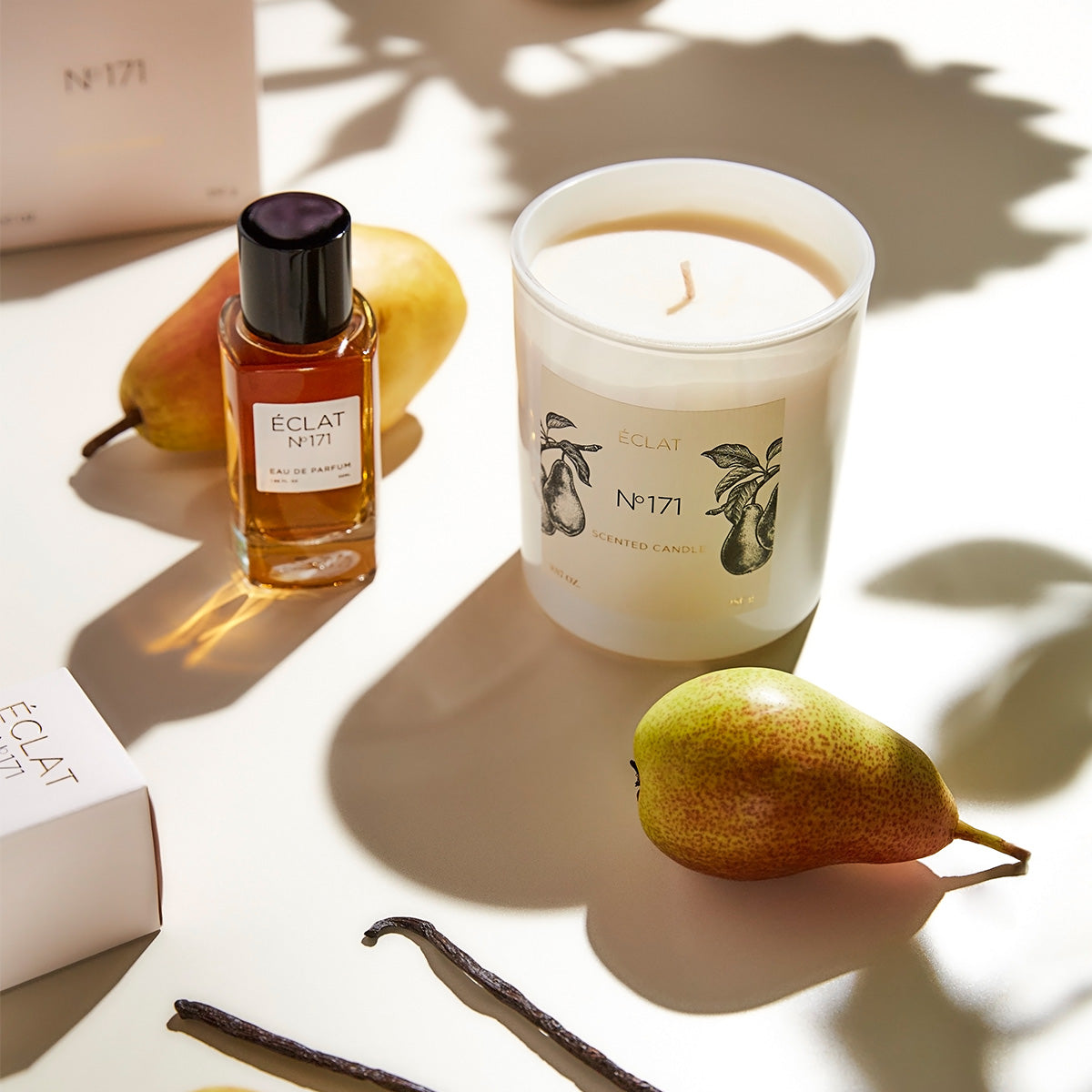 Glass bottle of ÉCLAT Blackcurrant & Praline perfume with black cap beside an elegant white candle and pear on a light background.