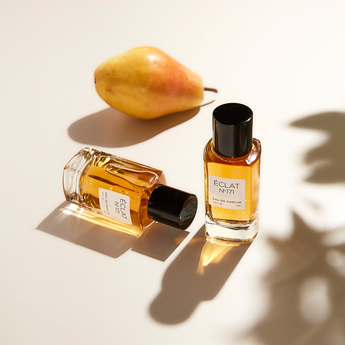 ÉCLAT Blackcurrant & Praline in elegant glass bottles with black caps, placed beside a ripe pear, casting soft shadows.