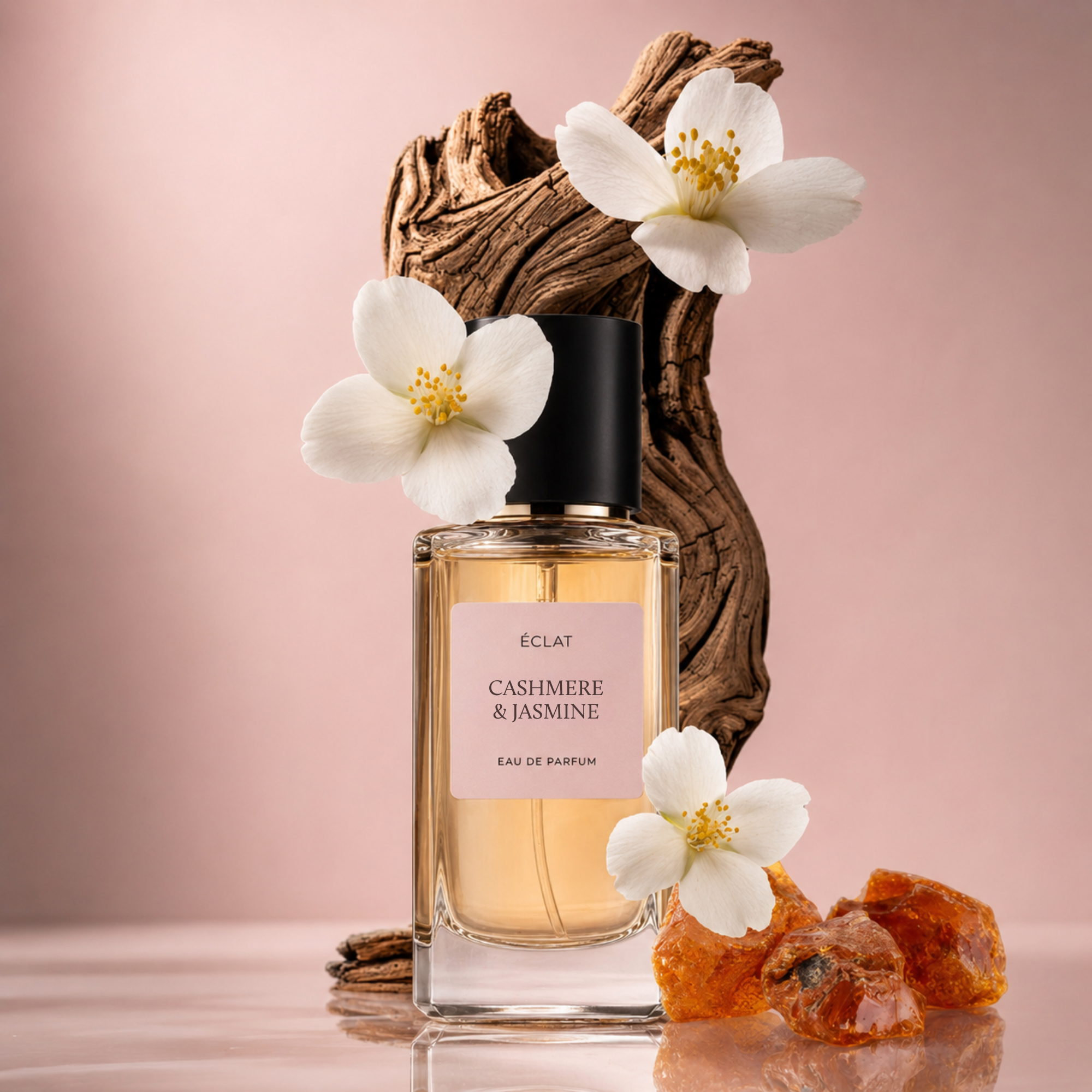A clear glass bottle of ÉCLAT Cashmere & Jasmine perfume with a black cap, surrounded by white jasmine flowers and amber stones on a textured wooden background.