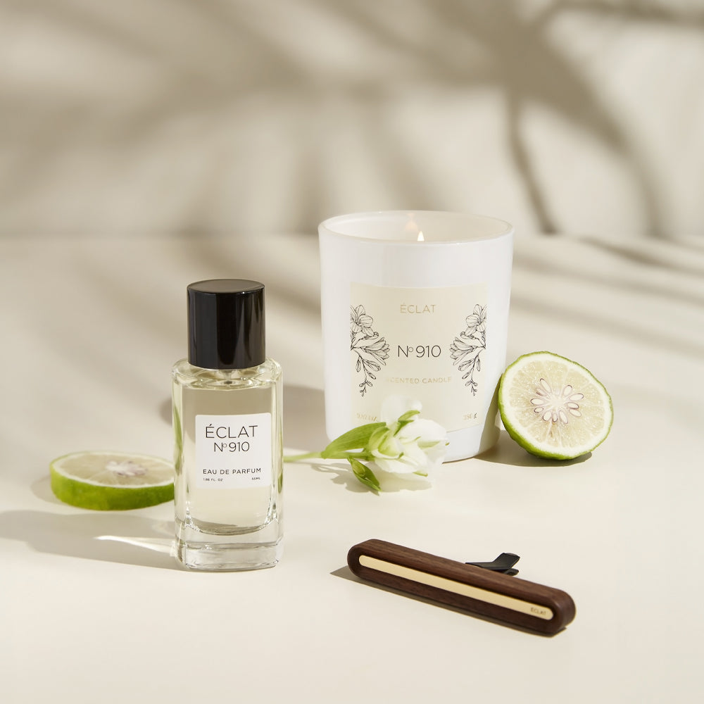 A faceted glass bottle of ÉCLAT Creamy White perfume with a black cap, beside a white scented candle, citrus slice, and flower.