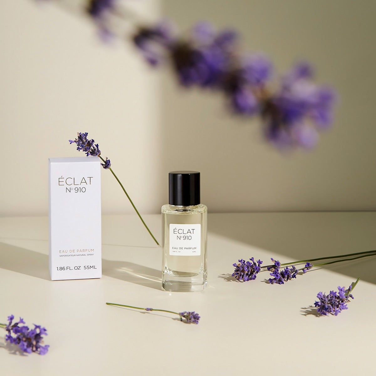 ÉCLAT Creamy White perfume in a clear glass bottle with a black cap, surrounded by lavender sprigs, stands beside its elegant white box.