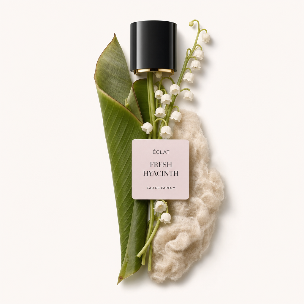 Une composition de feuilles vertes fraîches et de délicates fleurs blanches disposées aux côtés d'un matériau fibreux doux, surmontée d'un capuchon noir avec une étiquette ÉCLAT Fresh Hyacinth.