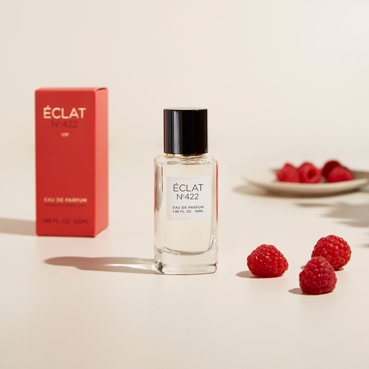 Flacon en verre transparent d'ÉCLAT Musky Passionfruit avec un bouchon noir, accompagné d'une boîte rouge et de framboises fraîches.