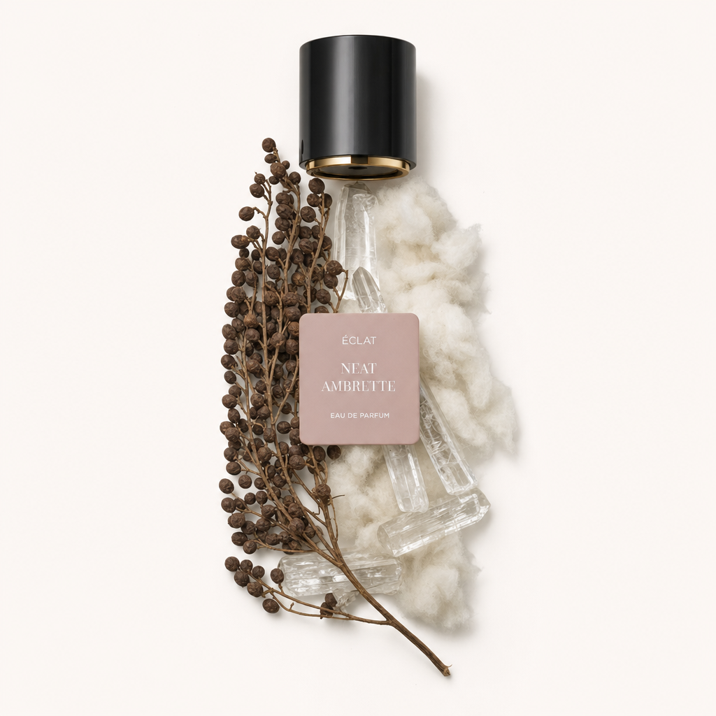 Un arrangement soigné de graines brunes et de coton blanc doux et moelleux à côté de structures cristallines claires. En haut se trouve un bouchon noir avec une étiquette beige portant ÉCLAT Neat Ambrette.
