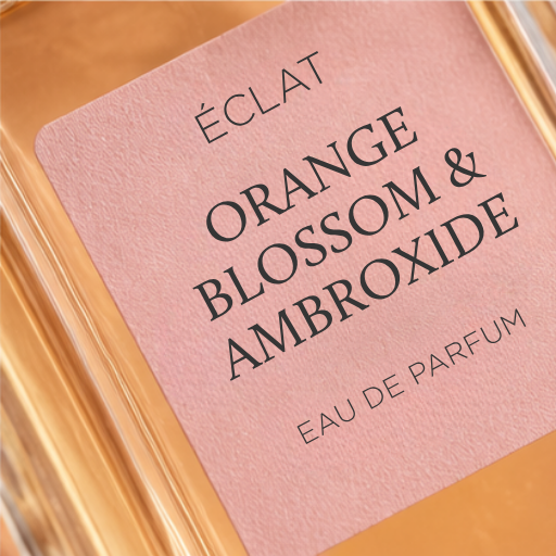 A faceted glass bottle with a pink label displaying the name ÉCLAT Orange Blossom & Ambroxide.
