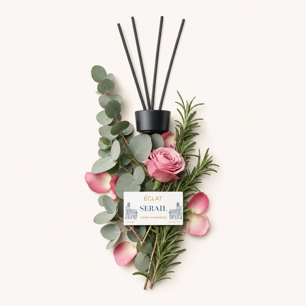 Uma composição de folhas de eucalipto, ramos de alecrim, pétalas de rosa rosa e uma rosa em flor rodeia uma tampa preta com etiqueta para ÉCLAT Serail Room Fragrance Sticks.