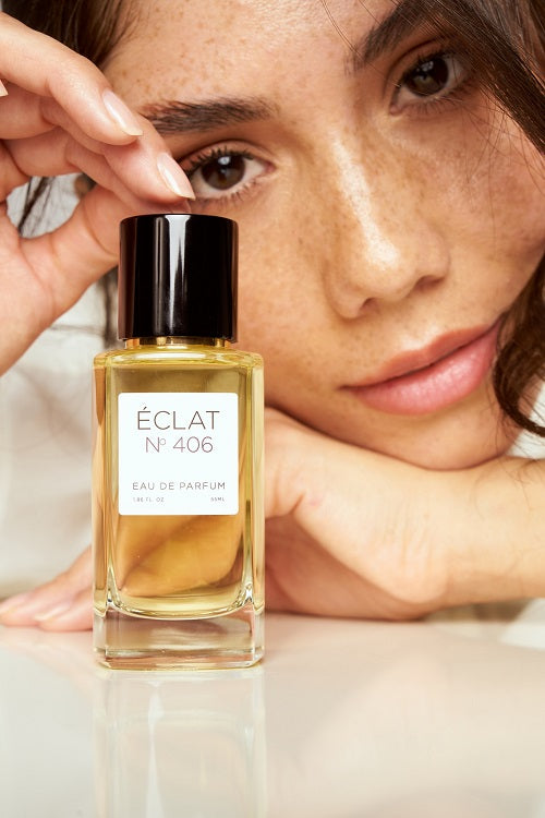 Un flacon en verre facetté de l'ÉCLAT N° 406 Eau de Parfum avec un bouchon noir, devant le visage d'une personne.