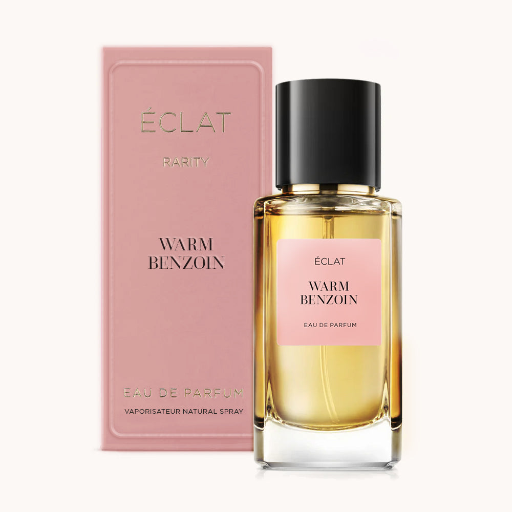 El perfume ÉCLAT Warm Benzoin se presenta con un frasco de vidrio transparente con tapa negra y etiqueta rosa; al lado hay una caja rosa a juego.