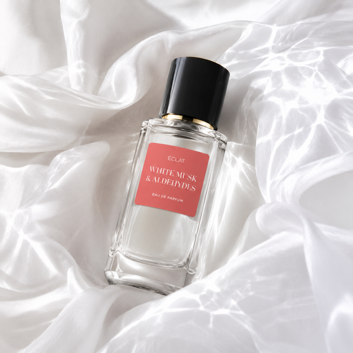 A faceted glass bottle of ÉCLAT White Musk & Aldehydes, featuring a black cap and coral label, rests on a satin fabric backdrop.